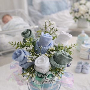 May include: A bouquet of baby socks arranged to resemble flowers, in shades of blue, green, and white. The arrangement is in a small, striped bucket, with greenery and small white flowers. A thoughtful gift for a new baby.