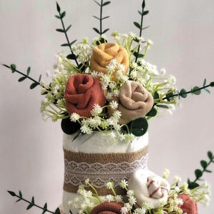 May include: A three-tiered diaper cake constructed from white rolled diapers, decorated with burlap and white lace ribbons. The tiers are adorned with fabric flowers rolled from socks in shades of mustard, rust, and cream, accented with sprigs of greenery.