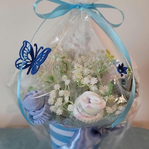 May include: A baby gift basket wrapped in clear cellophane and tied with a light blue ribbon. The basket contains white flowers, rolled baby socks, and a blue butterfly decoration. The container is a blue and white striped cup.