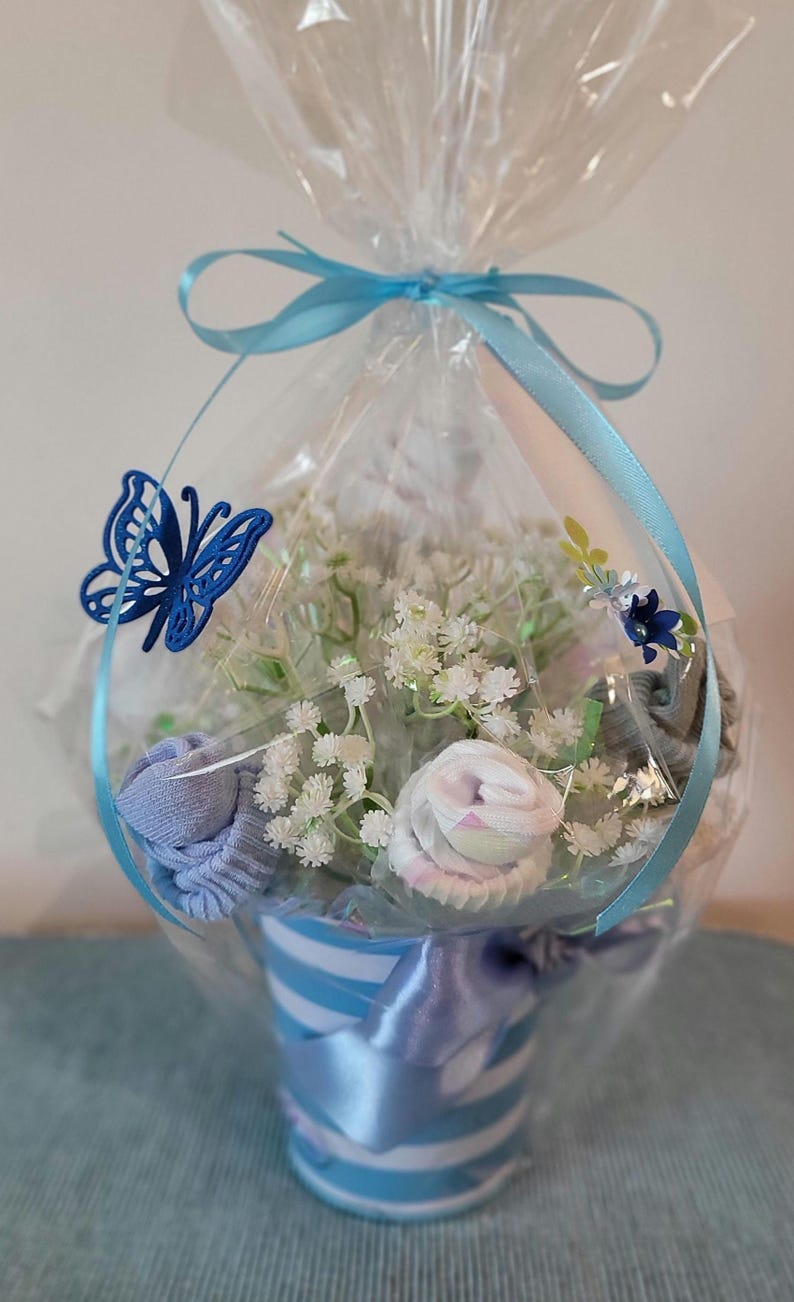 May include: A baby gift basket wrapped in clear cellophane and tied with a light blue ribbon. The basket contains white flowers, rolled baby socks, and a blue butterfly decoration. The container is a blue and white striped cup.