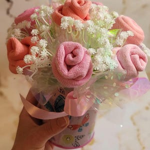 May include: A bouquet of pink and peach-colored sock roses, accented with white filler flowers, presented in a decorative container. The container is wrapped with a pink ribbon and iridescent cellophane.