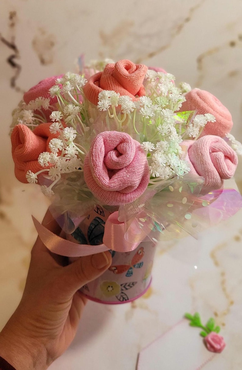 May include: A bouquet of pink and peach-colored sock roses, accented with white filler flowers, presented in a decorative container. The container is wrapped with a pink ribbon and iridescent cellophane.