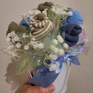 May include: A bouquet of socks arranged to resemble flowers, in a white bucket with a blue ribbon. The sock flowers are various shades of green and blue, with white baby's breath and iridescent cellophane.
