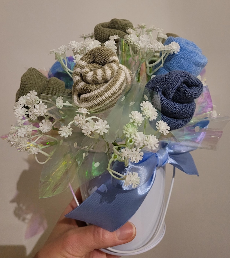May include: A bouquet of socks arranged to resemble flowers, in a white bucket with a blue ribbon. The sock flowers are various shades of green and blue, with white baby's breath and iridescent cellophane.