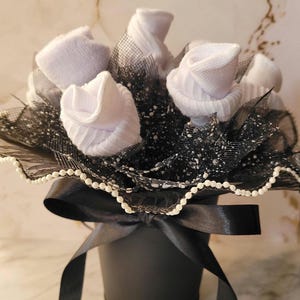 May include: A black bucket-shaped container holds a bouquet of white sock roses. The arrangement includes black netting and a black satin ribbon tied in a bow. The rim is trimmed with small white beads.