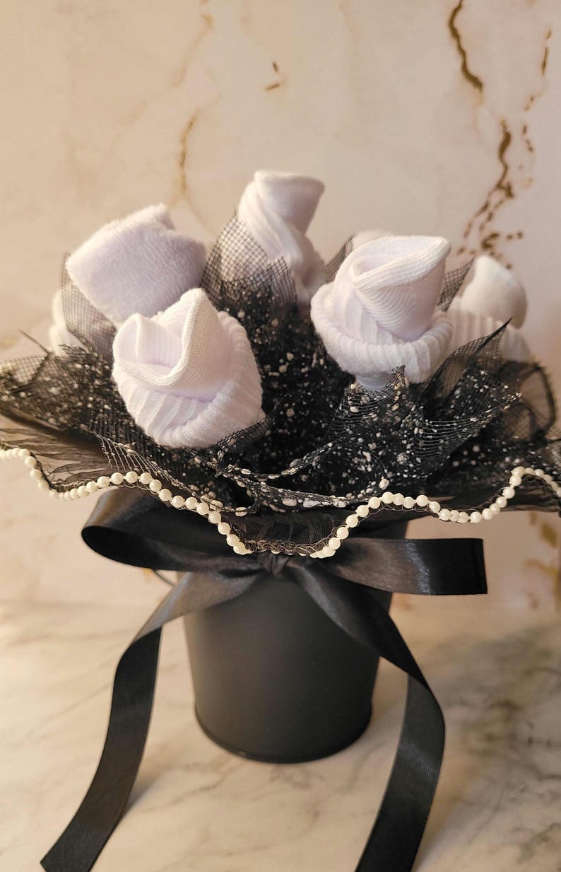 May include: A black bucket-shaped container holds a bouquet of white sock roses. The arrangement includes black netting and a black satin ribbon tied in a bow. The rim is trimmed with small white beads.