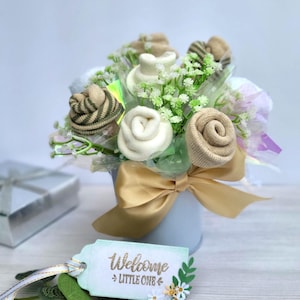 May include: A baby gift bouquet featuring sock roses in shades of beige, white, and green, arranged with faux greenery in a light blue pot. A gold ribbon and a tag that says "Welcome Little One" complete the arrangement.