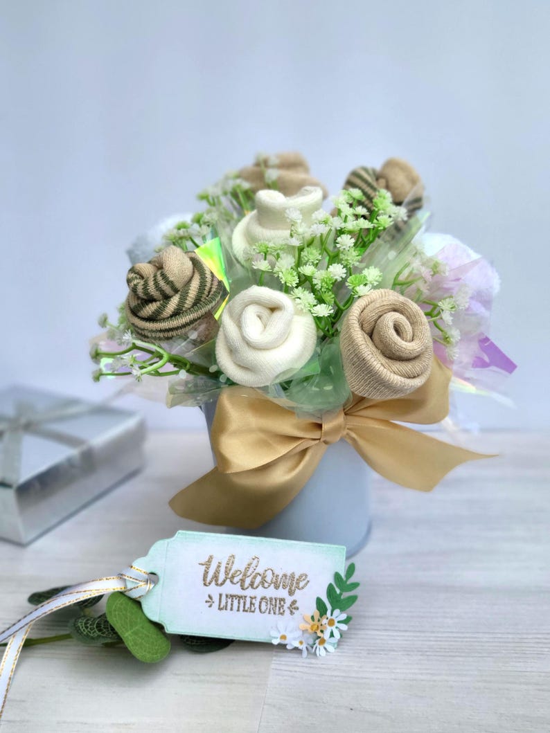 May include: A baby gift bouquet featuring sock roses in shades of beige, white, and green, arranged with faux greenery in a light blue pot. A gold ribbon and a tag that says "Welcome Little One" complete the arrangement.