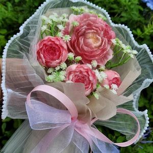 May include: A bouquet of pink peony-like flowers and small white flowers wrapped in tan paper and white lace. A pink ribbon is tied around the bouquet. The background is green.