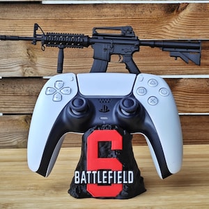 May include: A white and black PlayStation 5 controller with a black rifle on top. The controller rests on a black base with the number "6" in red and the word "BATTLEFIELD" in white.