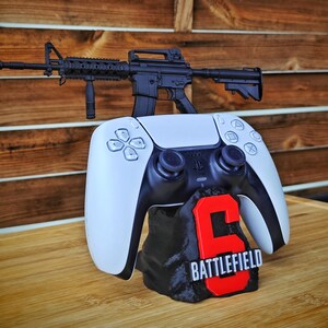 May include: A black and white PlayStation 5 controller with a black rifle on top. The controller rests on a black base with a red "6" and the word "BATTLEFIELD" in white.