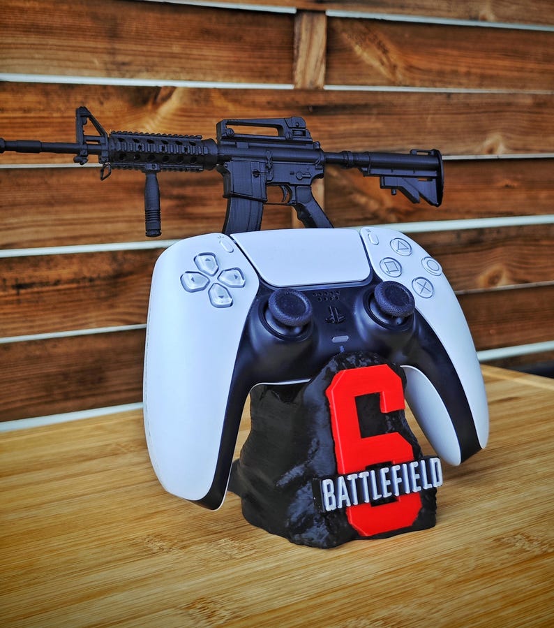 May include: A black and white PlayStation 5 controller with a black rifle on top. The controller rests on a black base with a red "6" and the word "BATTLEFIELD" in white.