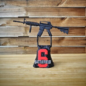 May include: A black rifle model is displayed above a black base with the number "6" in red and the word "BATTLEFIELD" below it. The background is a wooden wall.