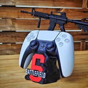May include: A white and black PlayStation 5 controller with a black and red "Battlefield 6" stand, and a black toy rifle. The controller is resting on the stand, with the rifle on top. The stand has the number "6" in red.