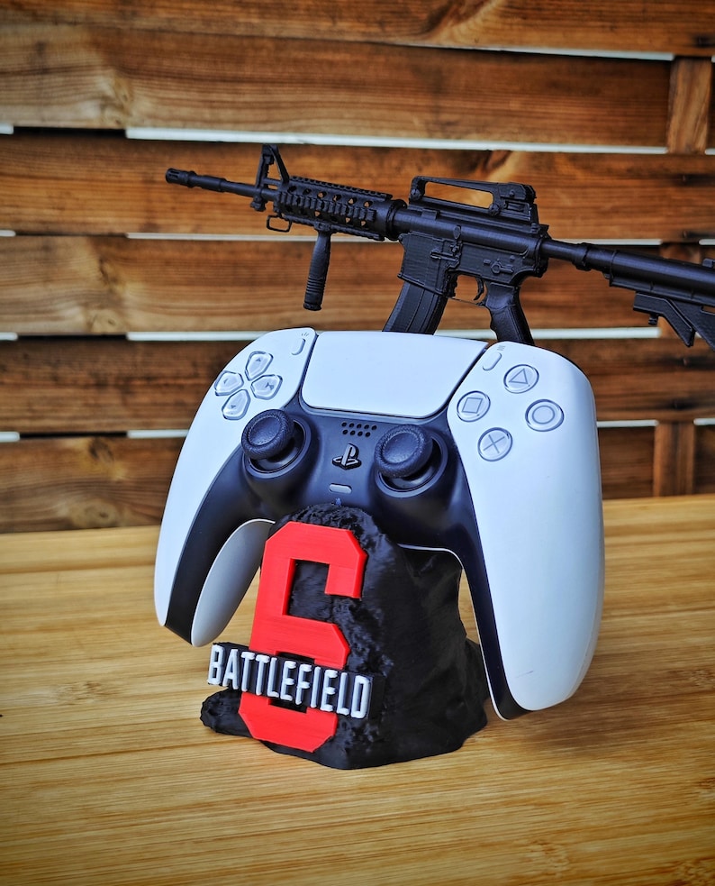 May include: A white and black PlayStation 5 controller with a black and red "Battlefield 6" stand, and a black toy rifle. The controller is resting on the stand, with the rifle on top. The stand has the number "6" in red.