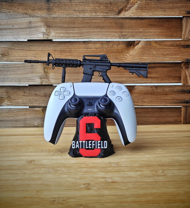 May include: A white and black PlayStation 5 controller with a black rifle on top. The controller rests on a black base with the number "6" in red and the word "BATTLEFIELD" in white.