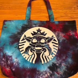 May include: A tie-dye tote bag with a white silhouette of a character holding a cup. The character is inside a white circle with a star on top. The circle is surrounded by a black and white ring.