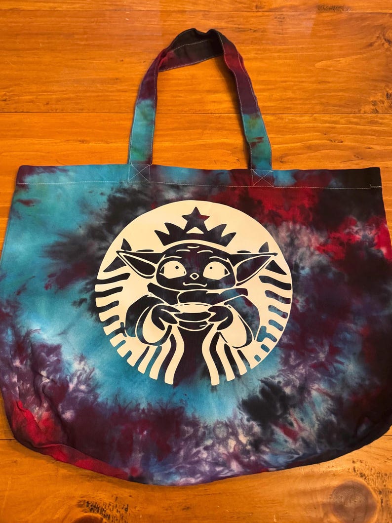 May include: A tie-dye tote bag with a white silhouette of a character holding a cup. The character is inside a white circle with a star on top. The circle is surrounded by a black and white ring.