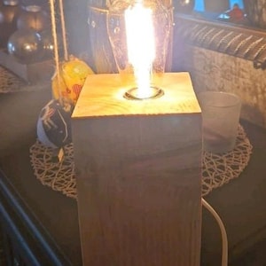 Puede incluir: Una base de lámpara de madera con una bombilla que brilla con una luz cálida. La base está hecha de madera natural con un acabado rústico.