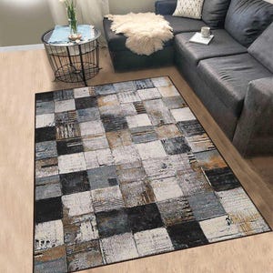 Puede incluir: Una alfombra rectangular con un diseño de patchwork. La alfombra presenta una mezcla de patrones cuadrados y rectangulares en tonos de negro, blanco, gris y beige. La alfombra está colocada sobre un suelo de madera clara, con un sofá seccional gris oscuro al fondo.