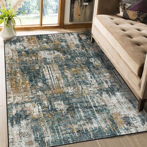 Puede incluir: Una alfombra rectangular con diseño abstracto. Presenta una mezcla de colores turquesa, dorado y blanco, creando una apariencia texturizada y desgastada. La alfombra está colocada sobre un suelo claro, cerca de un sofá beige y una ventana.