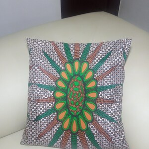 May include: Square cushion with a striking geometric design. The cushion showcases a central floral motif in green and orange, encircled by radiating brown and green lines against a white backdrop. The cushion is placed on a white surface.