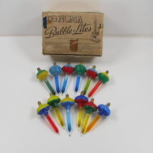May include: A set of 12 Noma Bubble-Lites Christmas tree lights with a cardboard box labeled 12 NOMA Bubble-Lites. The lights feature colorful plastic bases in red, blue, green, and yellow, with glass tubes containing liquid that bubbles when lit.