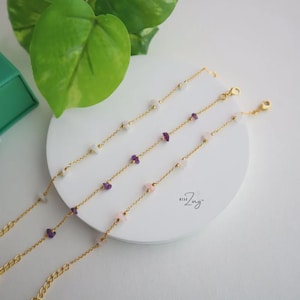May include: Three gold-toned chain bracelets featuring small, irregular gemstone chips in shades of white, purple, and light pink. Each bracelet includes a lobster claw clasp and a small extender chain. The text "Wear Zing" is printed on the white base.