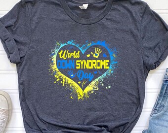 May include: A dark gray t-shirt with a blue and yellow heart design. The heart has the text "World Down Syndrome Day" inside. The t-shirt is paired with blue jeans and white sneakers.