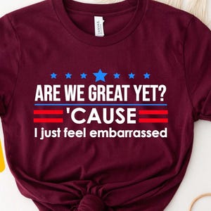 May include: Maroon t-shirt with the text "ARE WE GREAT YET? 'CAUSE I just feel embarrassed". The shirt has a red, white, and blue design with stars and stripes.