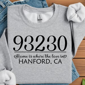 May include: A light gray sweatshirt with the number "93230" printed in large black letters. Below the number, the text reads "Home is where the love in HANFORD, CA". The sweatshirt is folded on top of blue jeans.