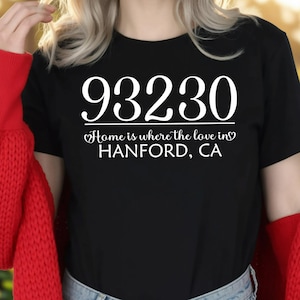 May include: Black t-shirt with the white text "93230 Home is where the love in HANFORD, CA." The shirt is paired with blue jeans and a red sweater. The background is a blurred outdoor scene with flowers.