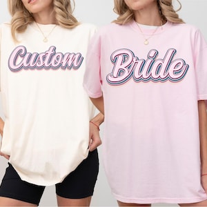 May include: Two oversized t-shirts, one ivory and one pink, each with a retro-style word graphic. The ivory shirt says "Custom" in a layered, colorful font. The pink shirt says "Bride" in a similar style. Both shirts have short sleeves.