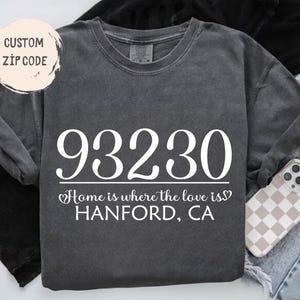 May include: A charcoal gray short-sleeve t-shirt featuring the white text 93230, followed by the phrase Home is where the love is and HANFORD, CA. A circular graphic in the top left corner displays the text CUSTOM ZIP CODE in black lettering.