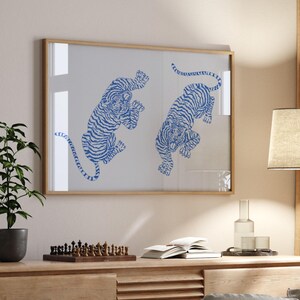 May include: A framed print of two blue and white stylised tigers on a white background. The frame is light wood and the print is hanging on a light beige wall.