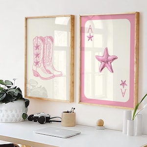 Puede incluir: Dos impresiones artísticas enmarcadas sobre un escritorio blanco. Una impresión presenta botas de vaquero rosas, y la otra muestra una estrella de mar rosa en una carta de juego. Los marcos son de madera clara.