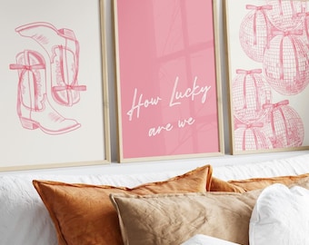 May include: Three framed prints with pink backgrounds and pink illustrations. The left print features a pair of pink cowboy boots. The middle print has the text "How lucky are we" in pink script. The right print features a pattern of pink disco balls.