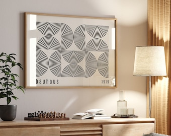 Puede incluir: Una impresión geométrica en blanco y negro enmarcada con el texto "bauhaus 1919" colgada en una pared blanca sobre una estantería de madera con un tablero de ajedrez, libros y una botella de vidrio. Un sofá gris con almohadas se encuentra frente a la estantería. Una lámpara de pie con una pantalla beige se encuentra junto al sofá.