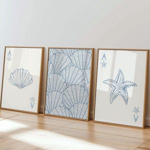 Pode incluir: Três impressões de arte emolduradas com tema costeiro. As impressões apresentam uma concha, um padrão de conchas e uma estrela do mar, todos em azul sobre fundo branco. Cada impressão tem uma moldura de madeira e o símbolo do Ás.