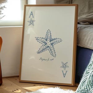 May include: Framed art print featuring a blue starfish design on a cream background. The artwork includes the text "Bonjour la mer" and the letter "A" with a starfish. The frame is made of light brown wood.