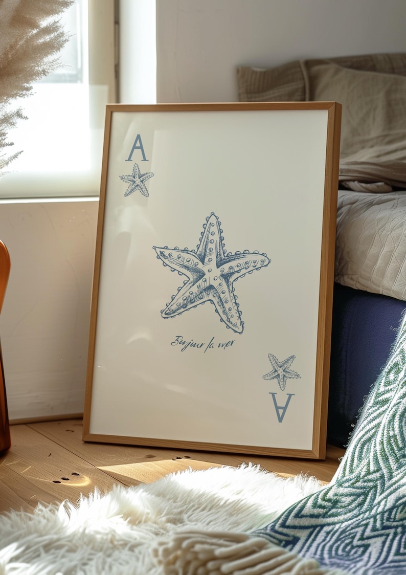 May include: Framed art print featuring a blue starfish design on a cream background. The artwork includes the text "Bonjour la mer" and the letter "A" with a starfish. The frame is made of light brown wood.