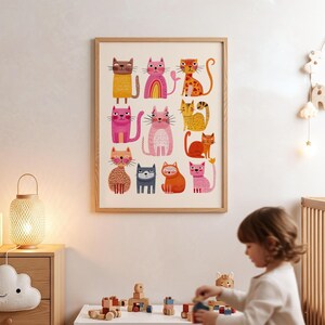 May include: A framed art print featuring a collection of colorful, cartoon-style cats. The cats are depicted in various poses and colors, including pink, orange, yellow, and brown. The artwork is displayed in a light-colored wooden frame, suitable for a child's room.