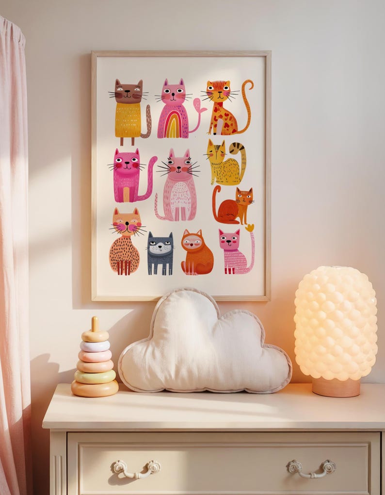 May include: A framed print featuring colorful cartoon cats in various poses. The artwork includes cats in shades of yellow, pink, orange, and blue. A wooden stacking toy, a cloud-shaped pillow, and a textured lamp sit on a white dresser.