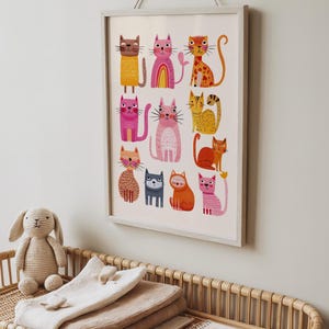 May include: A framed print featuring colorful, whimsical cat illustrations. The cats are depicted in various poses and colors, including pink, orange, and yellow. The artwork is displayed on a white background and framed in a light gray frame.