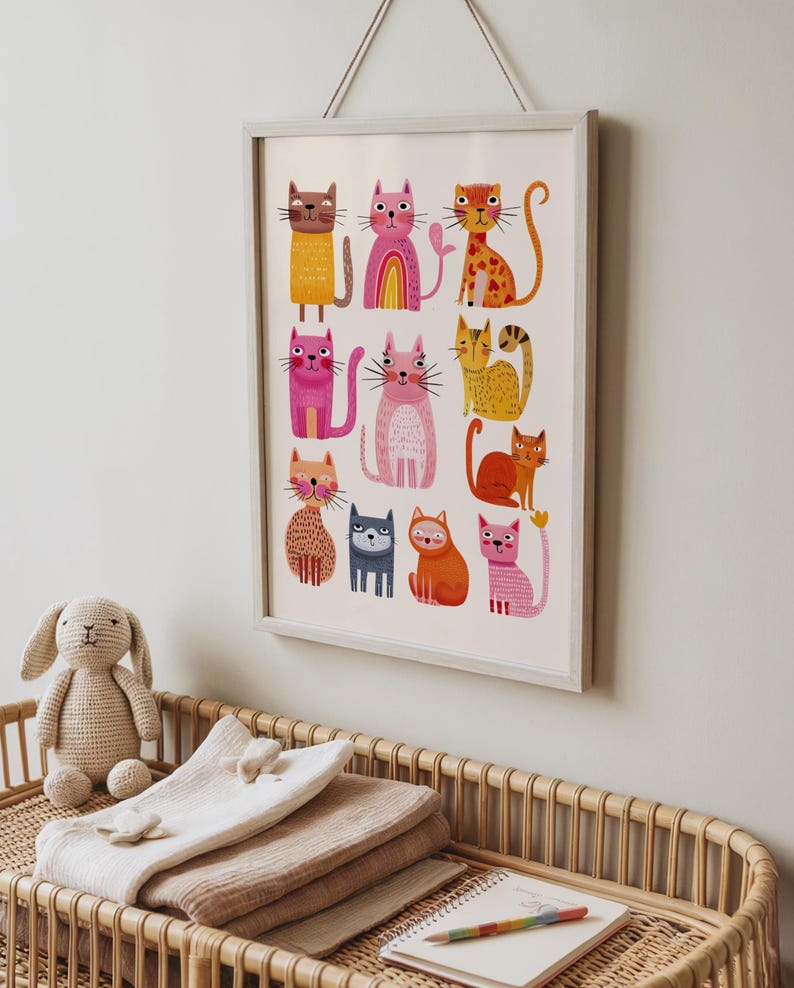 May include: A framed print featuring colorful, whimsical cat illustrations. The cats are depicted in various poses and colors, including pink, orange, and yellow. The artwork is displayed on a white background and framed in a light gray frame.