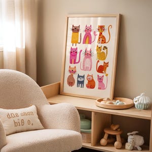 May include: A framed art print featuring colorful cat illustrations. The cats are depicted in various poses and colors, including orange, pink, and brown, with whimsical details. The artwork is displayed in a light-colored wooden frame.