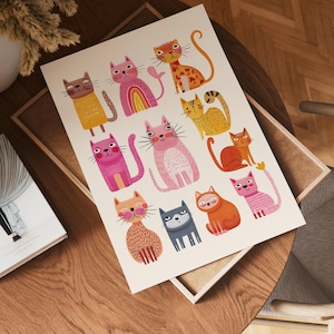 May include: A colorful art print featuring a collection of whimsical cat illustrations. The cats are depicted in various poses and colors, including pink, orange, yellow, and gray, with playful details like rainbow accents and heart shapes. The print is displayed on a wooden surface.