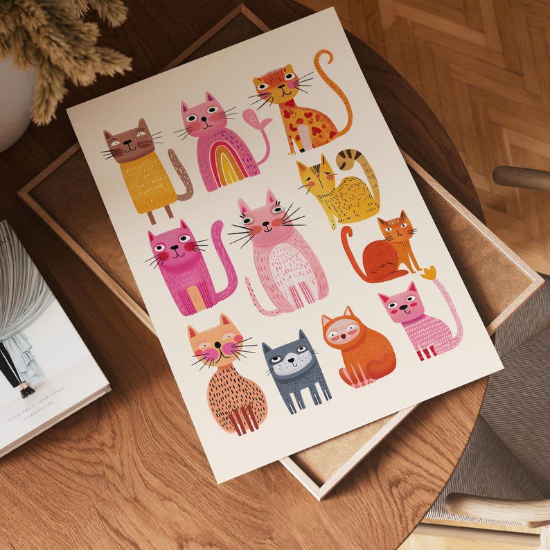May include: A colorful art print featuring a collection of whimsical cat illustrations. The cats are depicted in various poses and colors, including pink, orange, yellow, and gray, with playful details like rainbow accents and heart shapes. The print is displayed on a wooden surface.