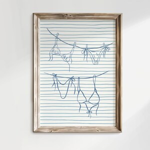 May include: A framed print featuring a line drawing of swimwear hanging on a clothesline. The illustration is in blue against a background of blue and white horizontal stripes. The wooden frame adds a rustic touch to the artwork.