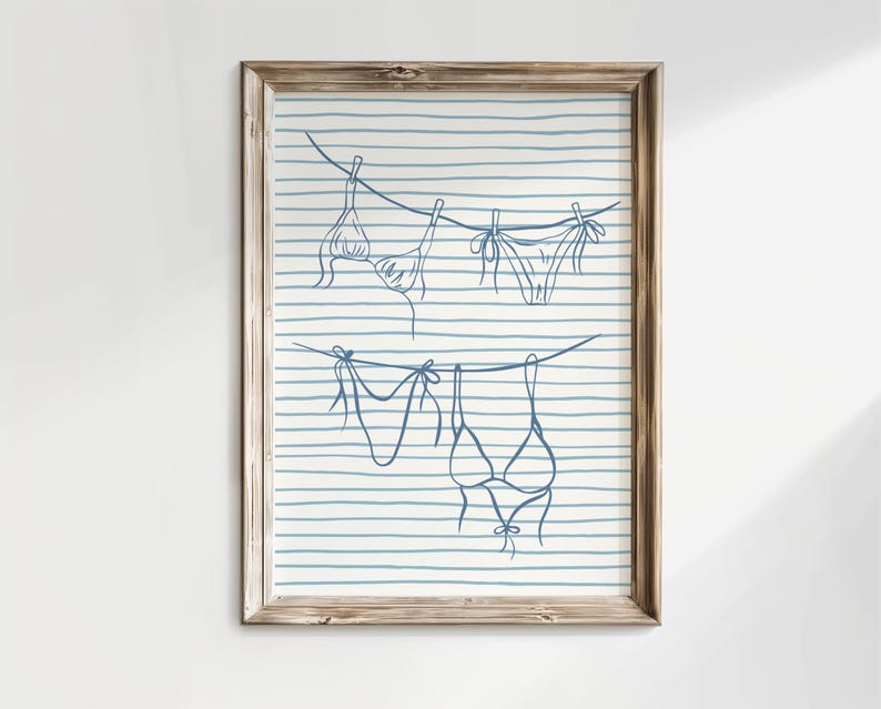 May include: A framed print featuring a line drawing of swimwear hanging on a clothesline. The illustration is in blue against a background of blue and white horizontal stripes. The wooden frame adds a rustic touch to the artwork.